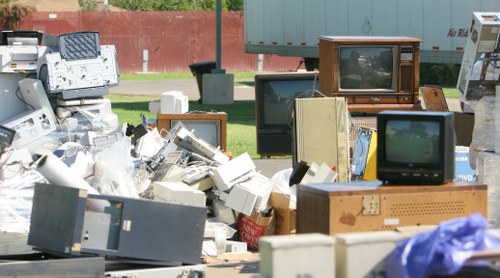 An outdoor scene featuring a collection of discarded electronic waste and old appliances arranged on a paved surface, with various vintage television sets in different sizes and models, some with wooden or plastic casings, visible in the foreground and middle ground. The televisions display static or blank screens, with some partially covered by plastic bags or cardboard boxes. To the left, additional electronic components and accessories are piled, including monitors, circuit boards, and miscellaneous device parts. In the background, a red brick wall and a green metal container or shed are visible, suggesting a designated waste or recycling area in Greenwich. The overall scene exhibits a mixture of textures, from the smooth plastic cases to the rougher wooden and metal surfaces, under natural daylight, emphasizing the importance of professional rubbish removal services such as those provided by Greenwich House Clearance to properly dispose of electronic waste in compliance with local environmental standards.