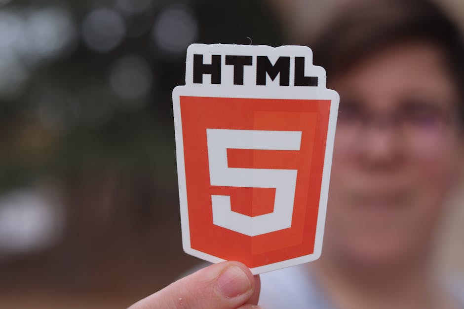 A person holding a small, rectangular sticker with a white border and a matte finish, displaying the HTML5 logo in the foreground, which consists of a bold, orange-shaded shield shape with a white num