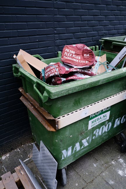 A green wheeled waste bin is placed on a paved surface beside a dark blue brick wall. The bin is filled with red and black bags of garden or landscaping materials, such as topsoil or mulch, marked wit