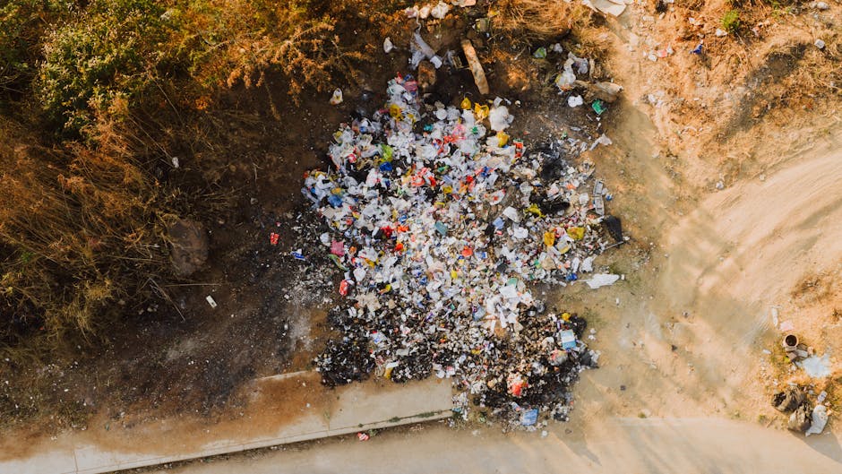 A green wheeled waste bin is placed on a paved surface beside a dark blue brick wall. The bin is filled with red and black bags of garden or landscaping materials, such as topsoil or mulch, marked wit